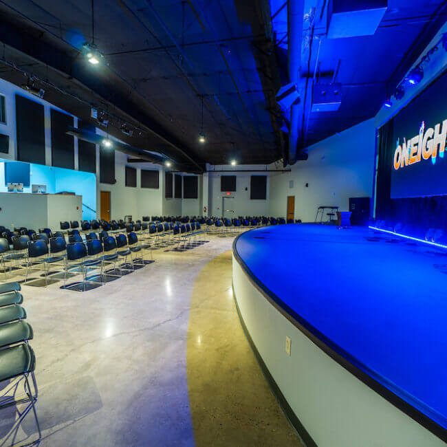 Interior of a modern auditorium with rows of empty black chairs and a large blue-lit stage. the word "oneighty" is displayed in bold on a screen above the stage.