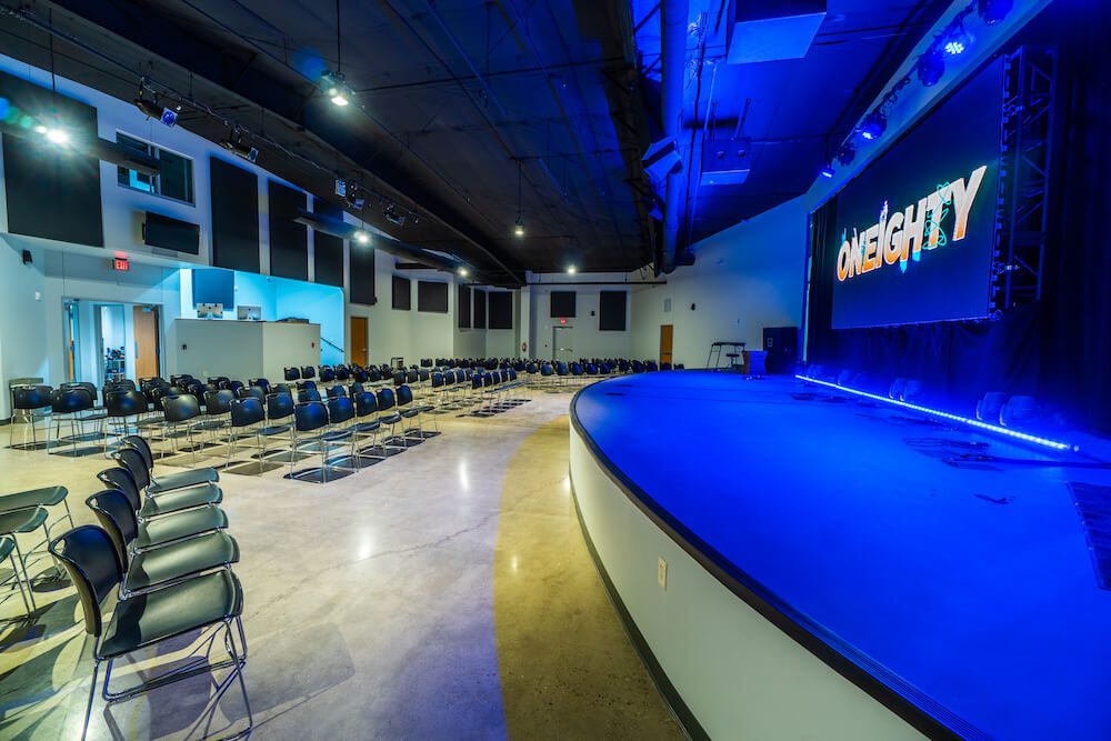 Interior of a modern auditorium with rows of empty black chairs and a large blue-lit stage. the word "oneighty" is displayed in bold on a screen above the stage.