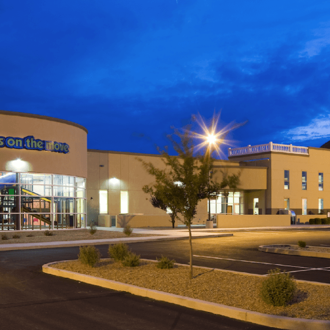 A modern church building at twilight, featuring a large sign "kids on the move" on the façade, a steeple, and an empty parking lot under a clear blue sky.