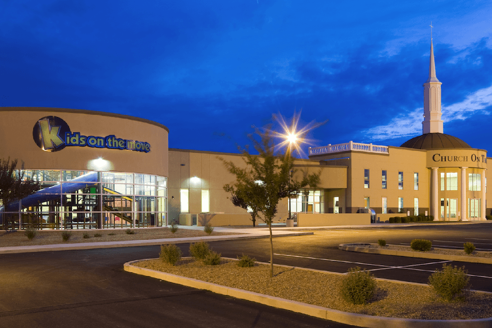A modern church building at twilight, featuring a large sign "kids on the move" on the façade, a steeple, and an empty parking lot under a clear blue sky.
