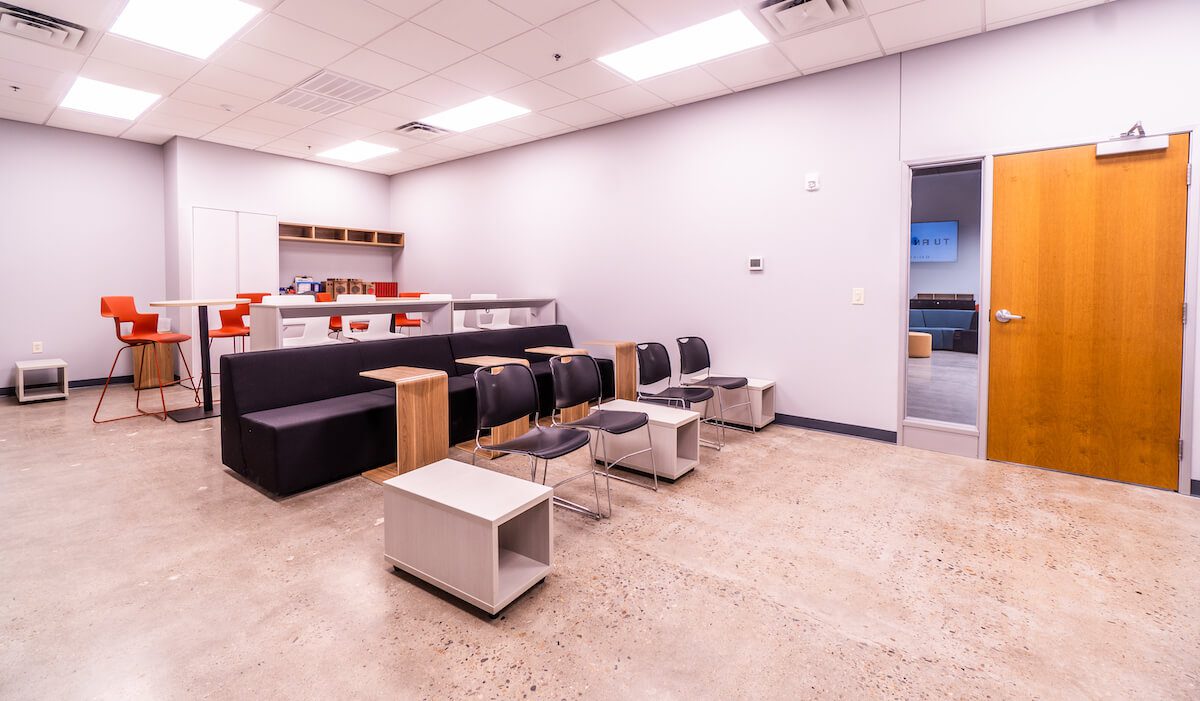 Modern office break room with a black sofa, several chairs, a coffee station designed by John Smith, and high stools at a counter, featuring neutral walls and a wooden door.