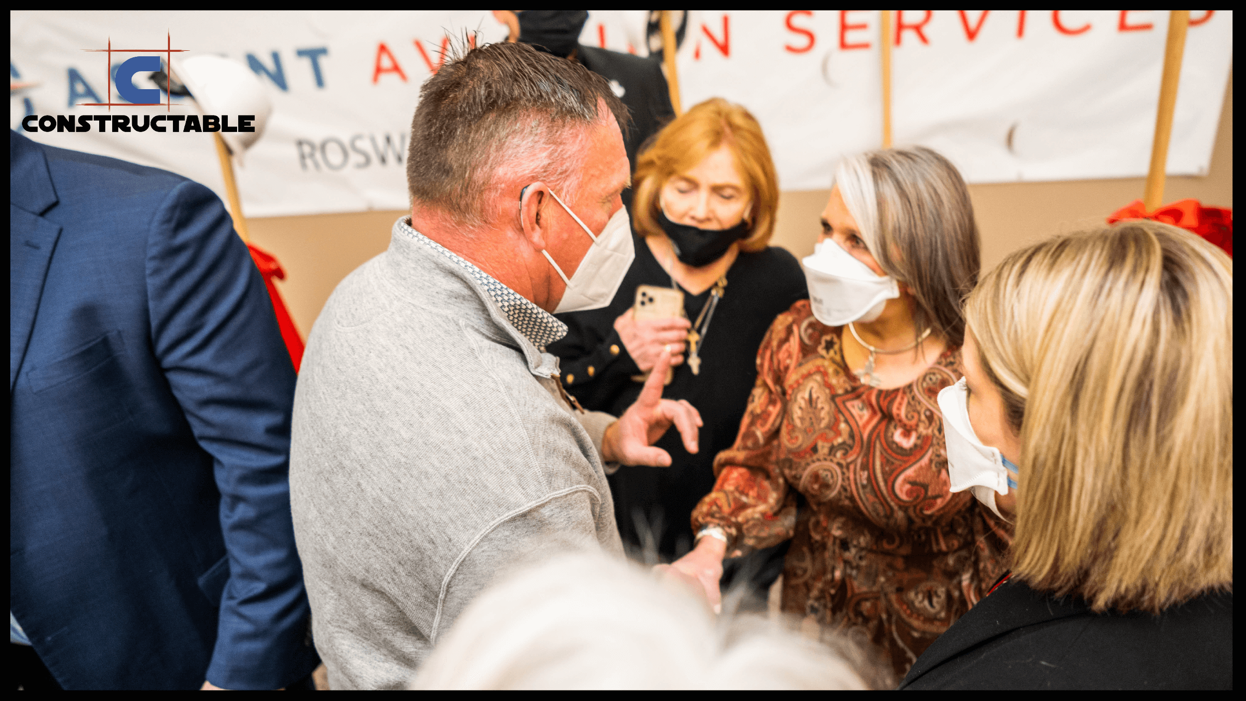 A group of adults, mostly wearing masks, engaged in a conversation at an indoor event with a banner in the background reading "construction costs annual service." The logo "constructable" is in the corner