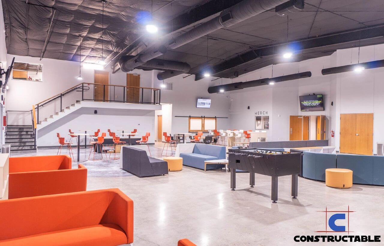 Modern office recreation area with orange couches, foosball and pool tables, arranged seating and desks, with a mezzanine level and the logo "construction costs" in the foreground.