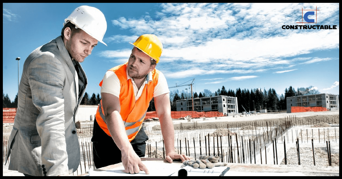 Two construction workers, one in a suit and another in a safety vest, review construction costs on a clipboard at a construction site with buildings in the background.