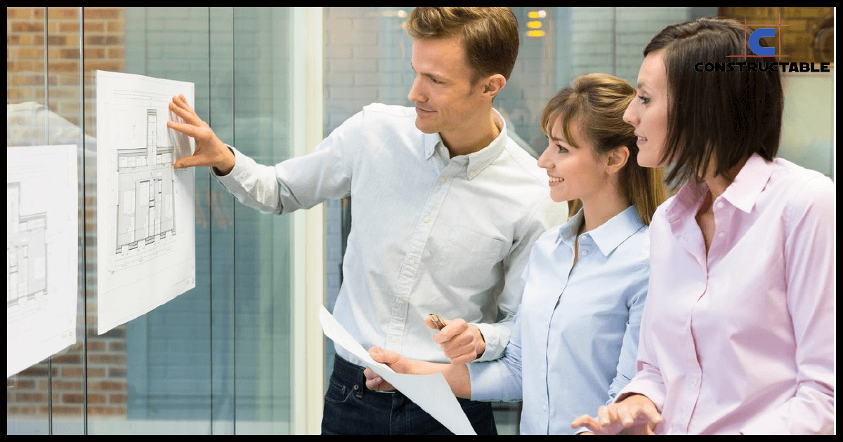 Three professionals reviewing architectural plans and construction costs pinned to a glass wall in an office setting, appearing engaged and focused on the task.