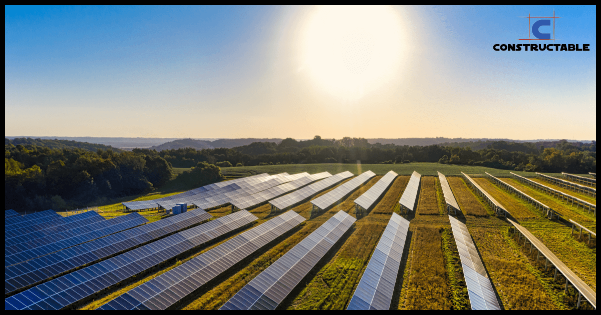 Aerial view of a vast solar farm with rows of solar panels gleaming under the bright sun, surrounded by lush greenery, with the horizon in the background and a clear sky overhead.