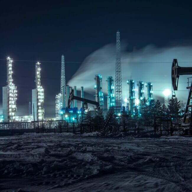 A nighttime view of an industrial area covered in snow, featuring illuminated oil rigs and refinery towers emitting steam under a dark, cloudy sky.