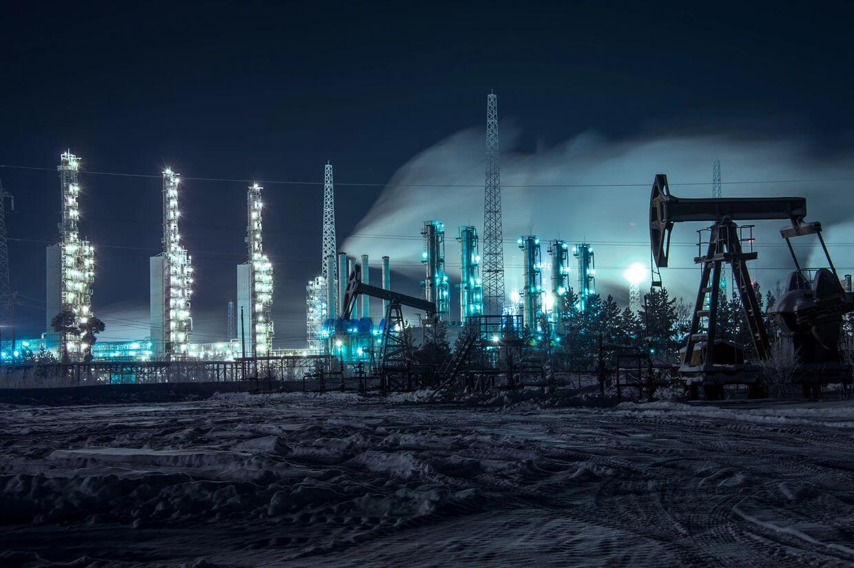 A nighttime view of an industrial area covered in snow, featuring illuminated oil rigs and refinery towers emitting steam under a dark, cloudy sky.