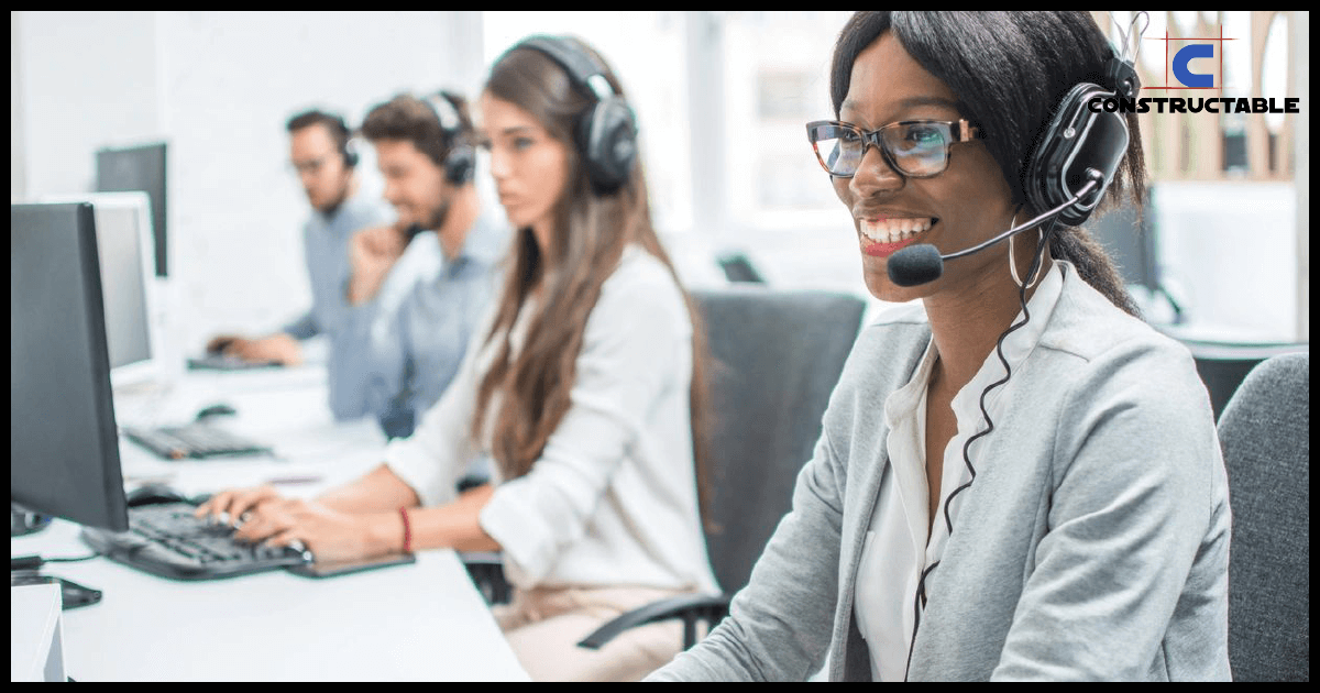 A diverse group of professionals wearing headsets and working at computers in a modern office environment, discussing construction costs. The focus is on a smiling woman in the foreground.