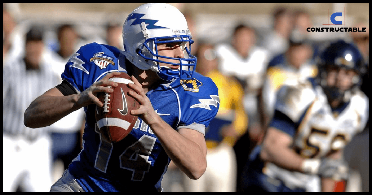 A football player in a blue and white uniform, wearing number 14, runs with the ball during a game, focusing intently as opponents from the yellow team approach, seemingly oblivious to rising construction costs