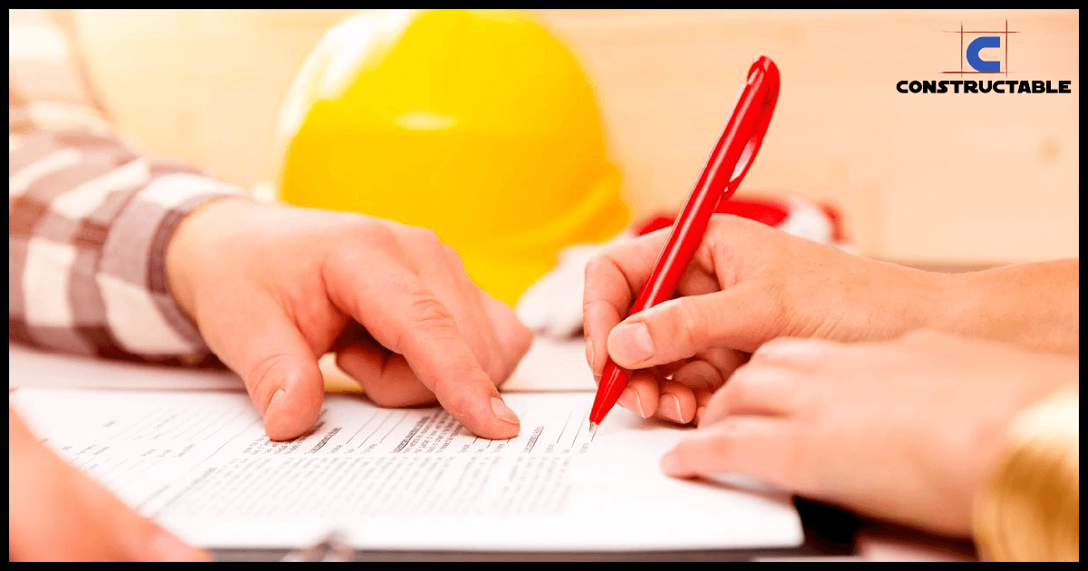 Two individuals, one wearing a plaid shirt, collaboratively review and annotate construction costs documents on a table, with a focus on a red pen and a yellow hard hat in the background.
