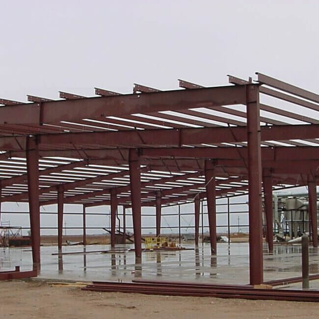 A partially constructed industrial steel building frame under a cloudy sky, with visible beams and columns on a concrete foundation.