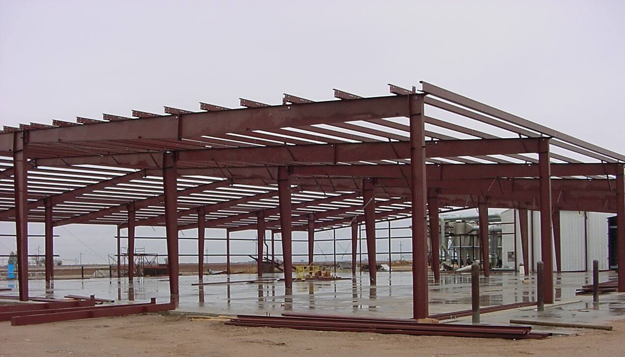 A partially constructed industrial steel building frame under a cloudy sky, with visible beams and columns on a concrete foundation.