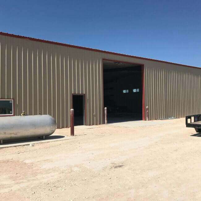 A large industrial warehouse with an open entrance, a metallic cylindrical tank on the left and a flatbed trailer on the right, under a clear blue sky.