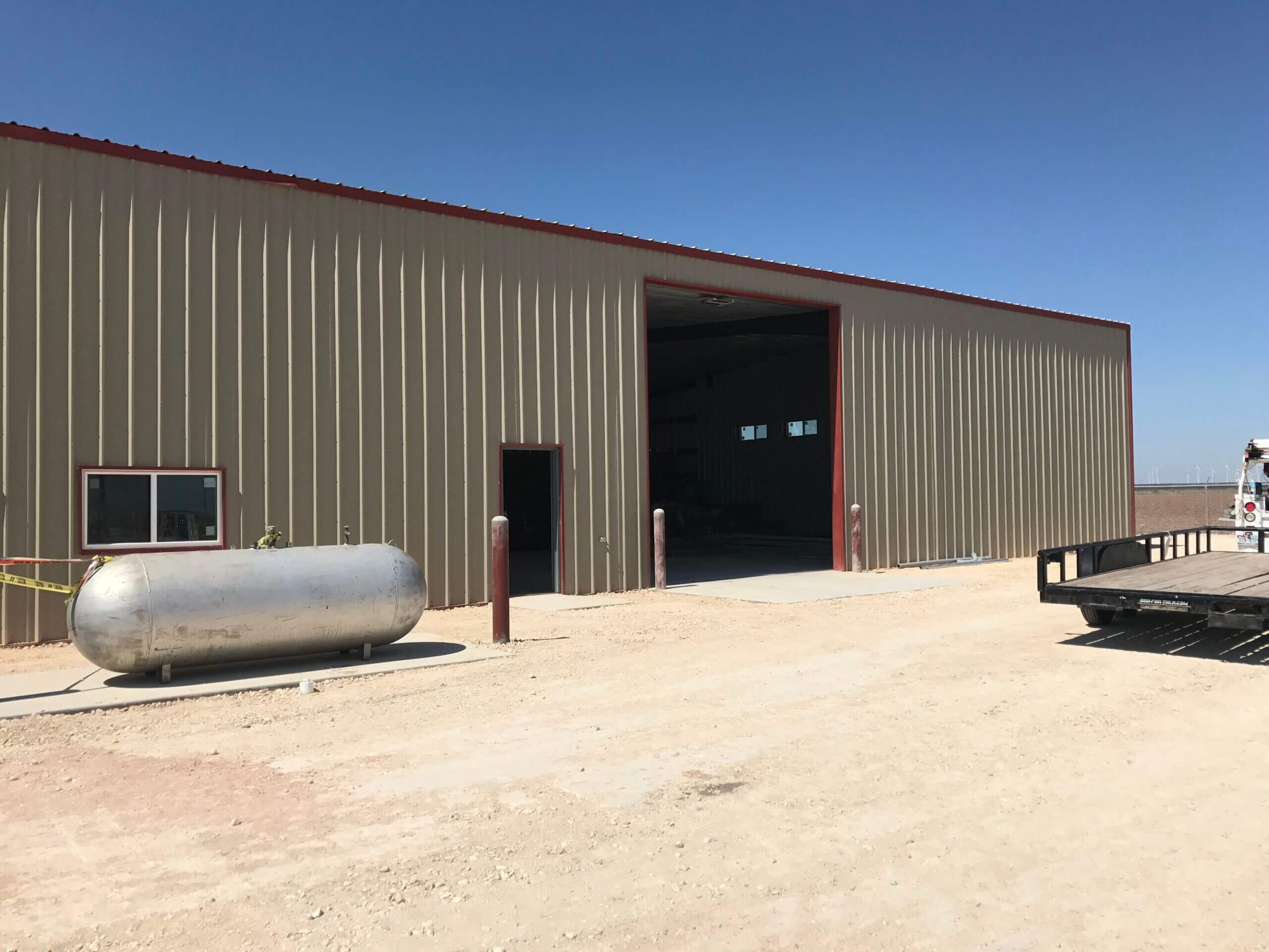 A large industrial warehouse with an open entrance, a metallic cylindrical tank on the left and a flatbed trailer on the right, under a clear blue sky.