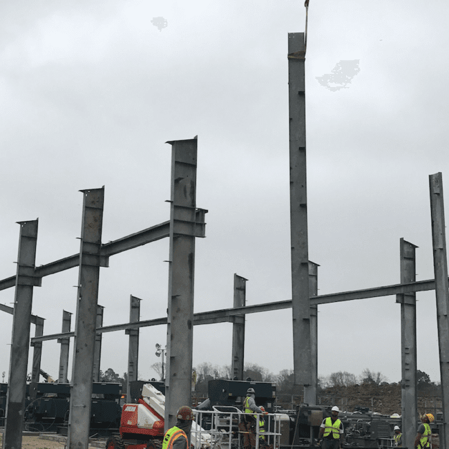 A construction site with steel columns being erected, forming the framework of a building, with construction workers and equipment visible under a cloudy sky.