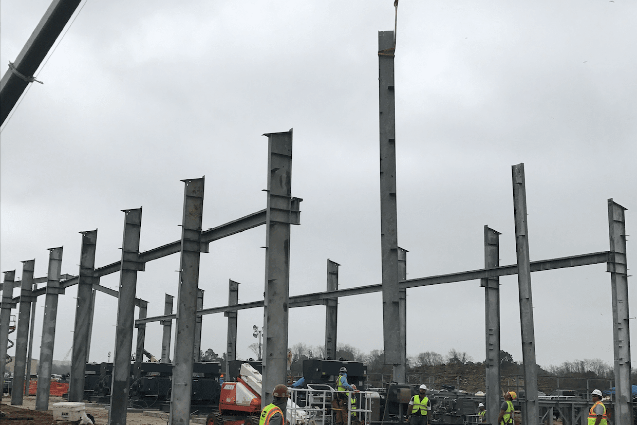A construction site with steel columns being erected, forming the framework of a building, with construction workers and equipment visible under a cloudy sky.
