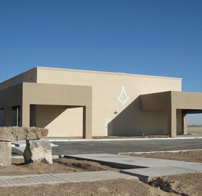 A modern beige single-story building with three garage doors, an american flag, and a masonic square and compass symbol on the facade, set in a sparsely landscaped area under a clear blue sky.