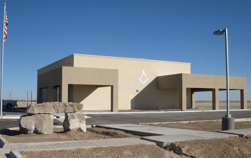 A modern beige single-story building with three garage doors, an american flag, and a masonic square and compass symbol on the facade, set in a sparsely landscaped area under a clear blue sky.