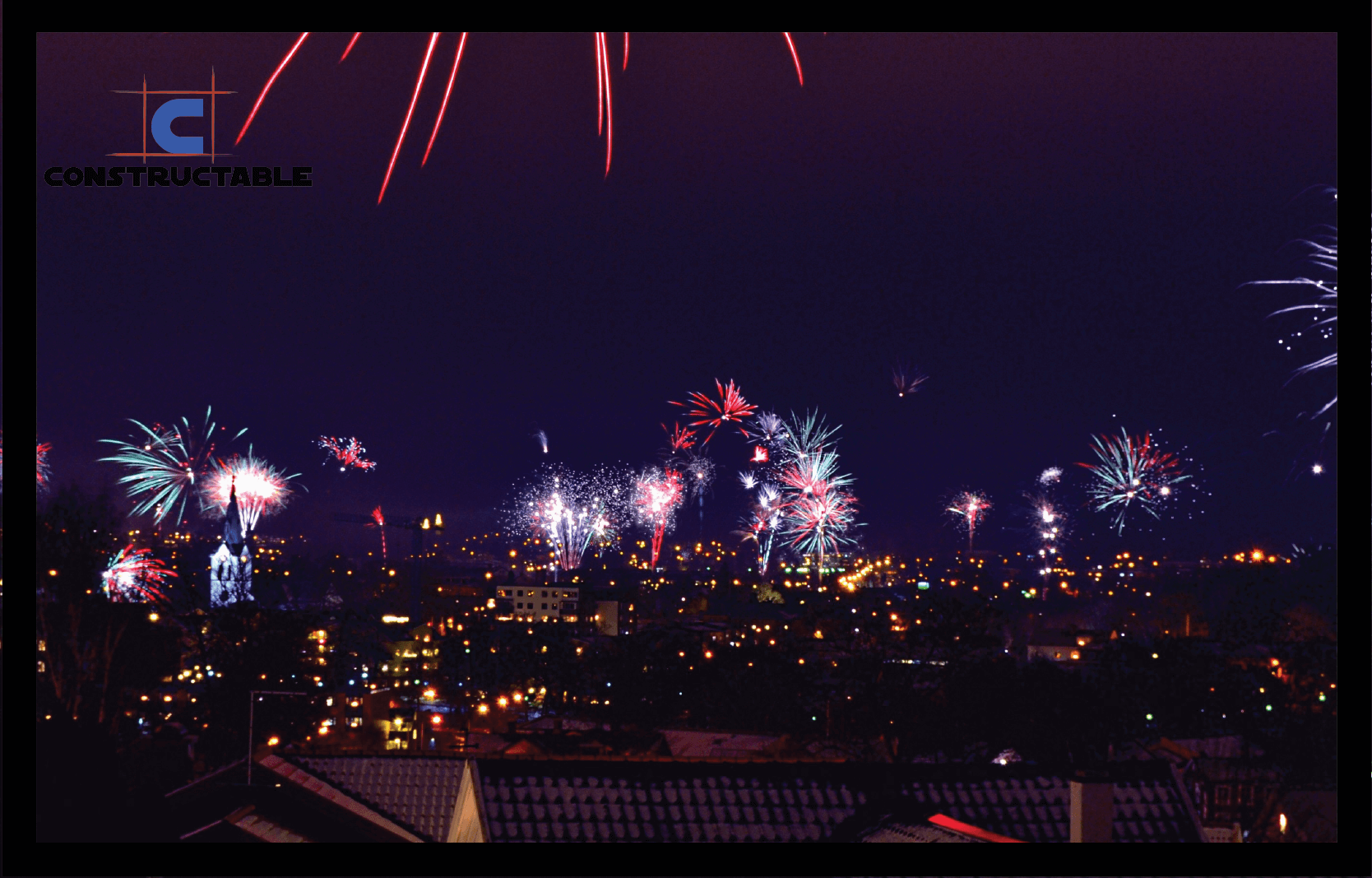 A vibrant display of multicolored fireworks bursts above a cityscape at night, with buildings silhouetted against the illuminated sky and a 2021 logo in the upper left corner.