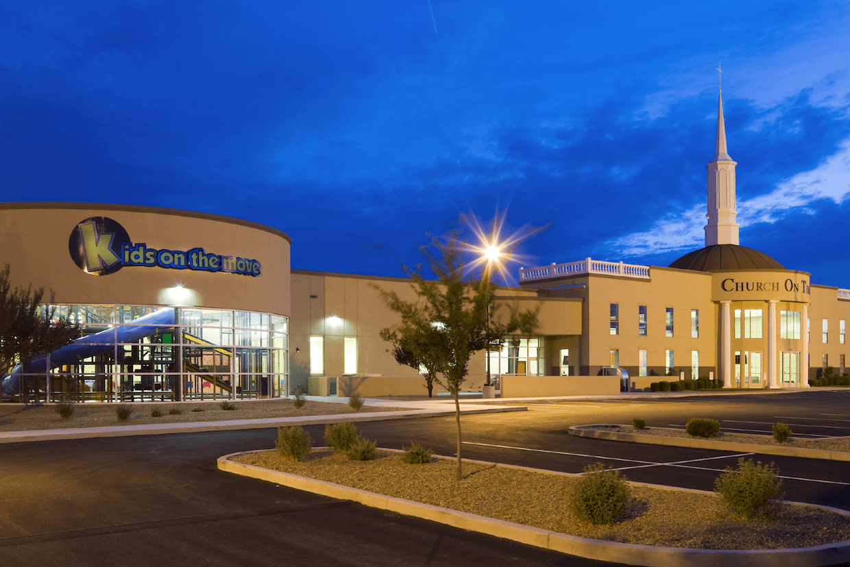 A modern church building at twilight featuring a curved front with "kids on the move" signage, an adjoining traditional steeple, and a clear blue sky. an empty parking lot is visible in the foreground.