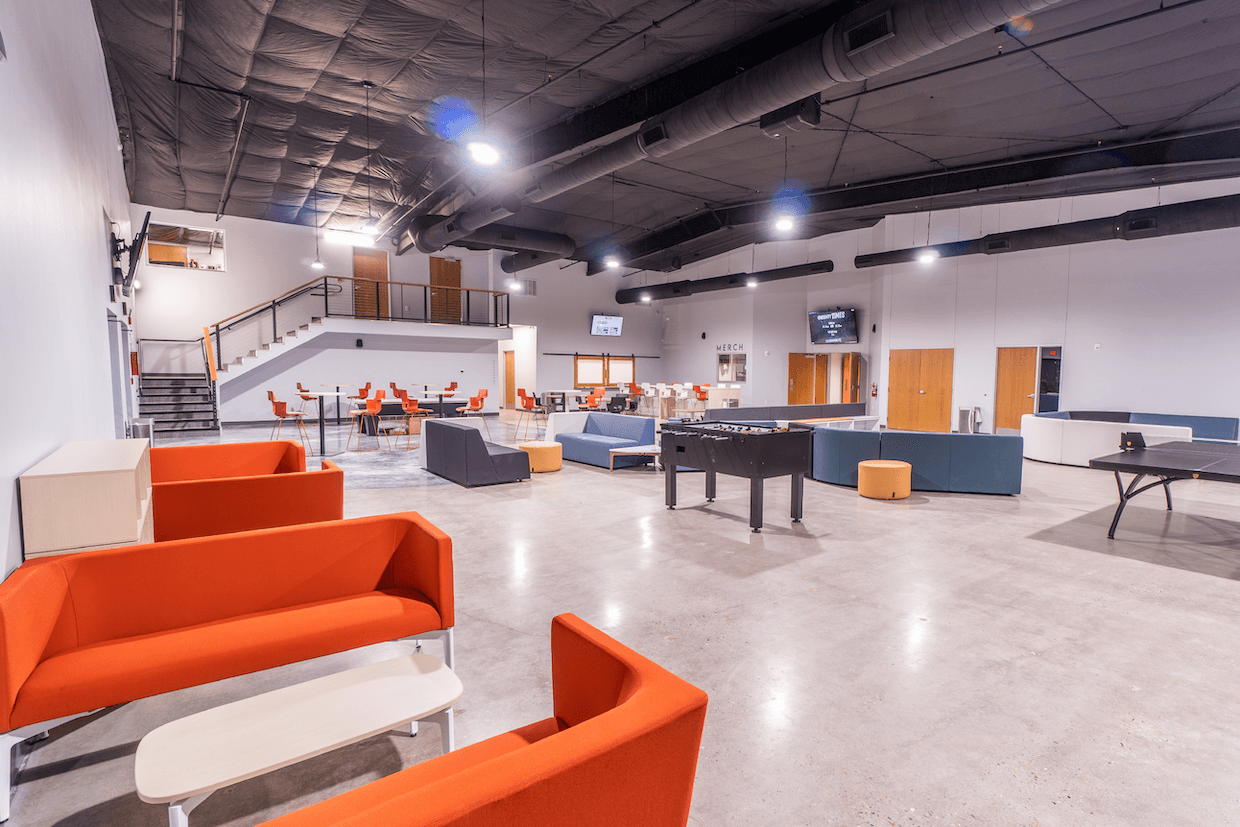 Modern spacious lounge with orange sofas, a ping pong table, coffee tables, and a dining area in the background with white chairs and tables. stairs lead to a loft space.