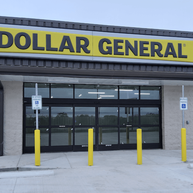 Exterior view of a dollar general store with a prominent yellow sign above the entrance. the building has a gray facade with glass doors and yellow protective bollards in front.