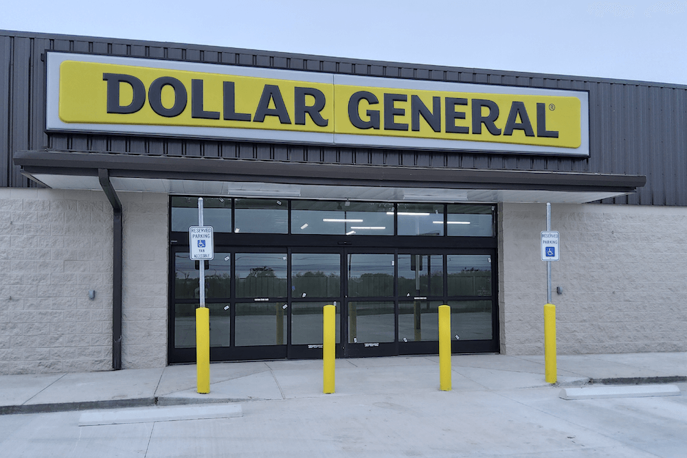 Exterior view of a dollar general store with a prominent yellow sign above the entrance. the building has a gray facade with glass doors and yellow protective bollards in front.