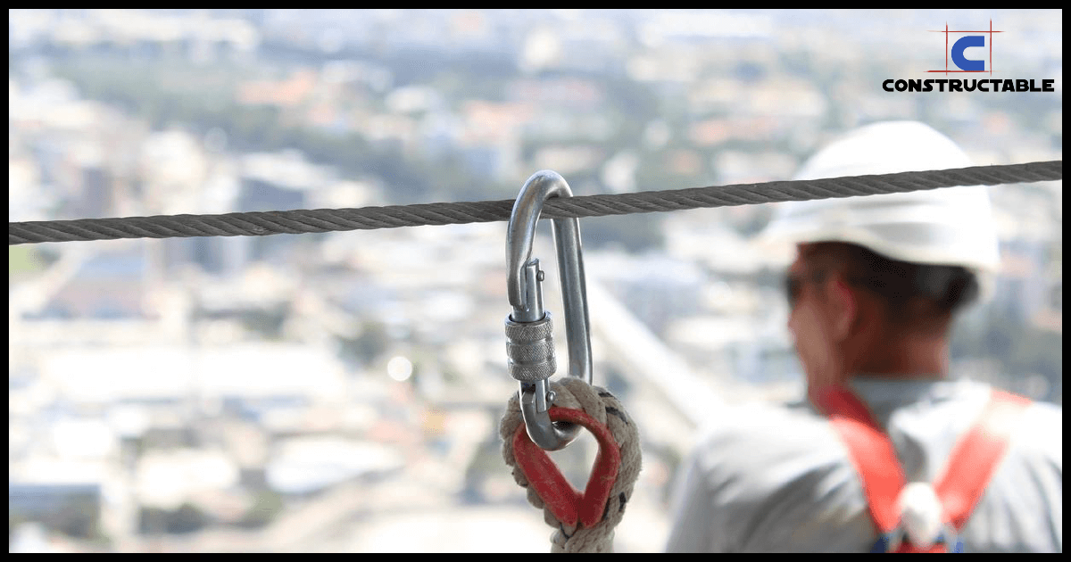 A construction worker in a hard hat and reflective vest looks out over a cityscape, contemplating construction costs. Seen out of focus in the background, in focus, a metal carabiner attaches to a