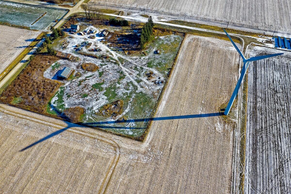 Aerial view of a rural landscape in winter showing fields with traces of snow, roads, a farm, and two large wind turbines casting long shadows on the ground.
