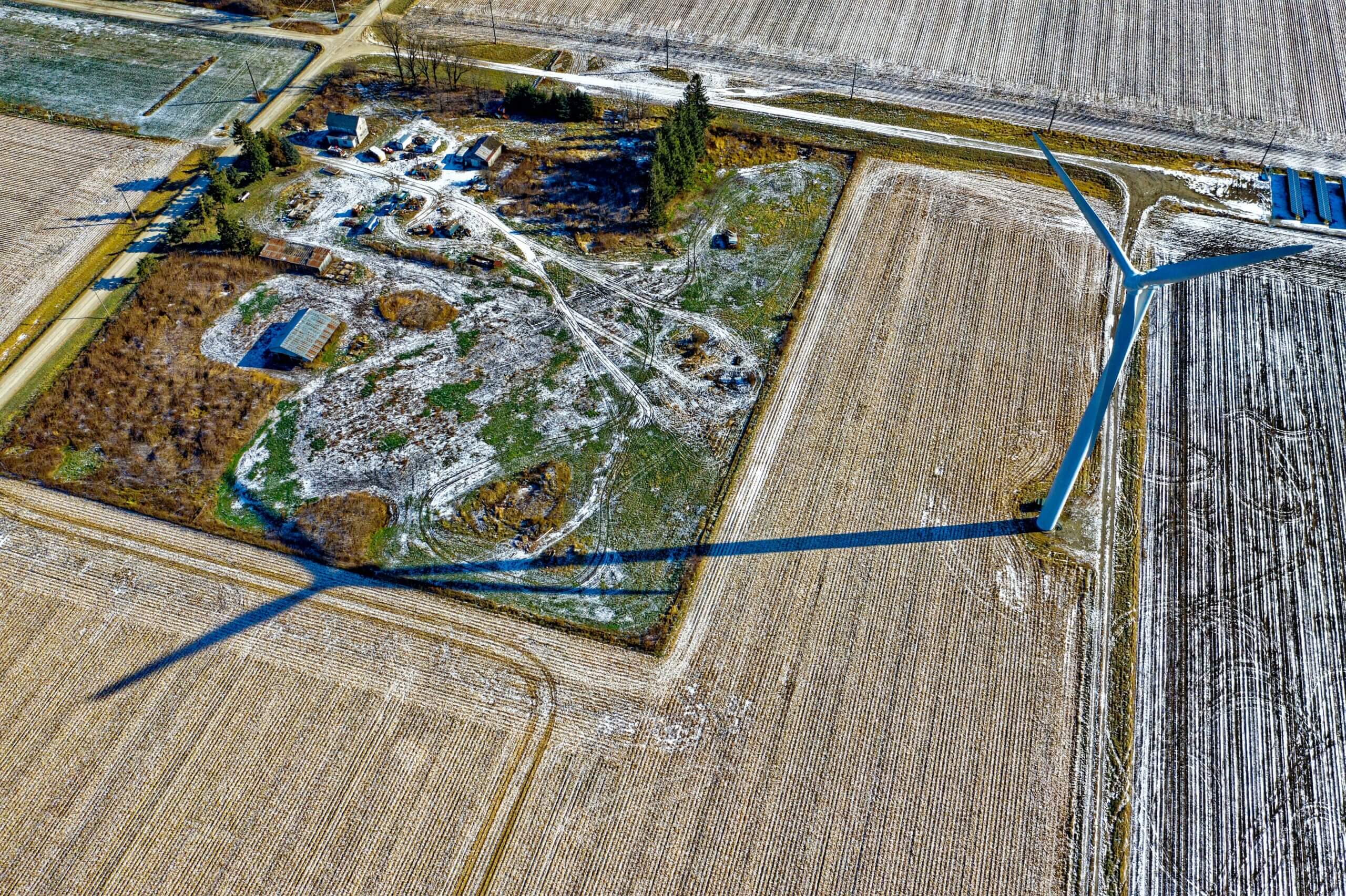 Aerial view of a wind farm casting long shadows across a wintery farmland with patches of snow, adjacent roads, and scattered buildings.