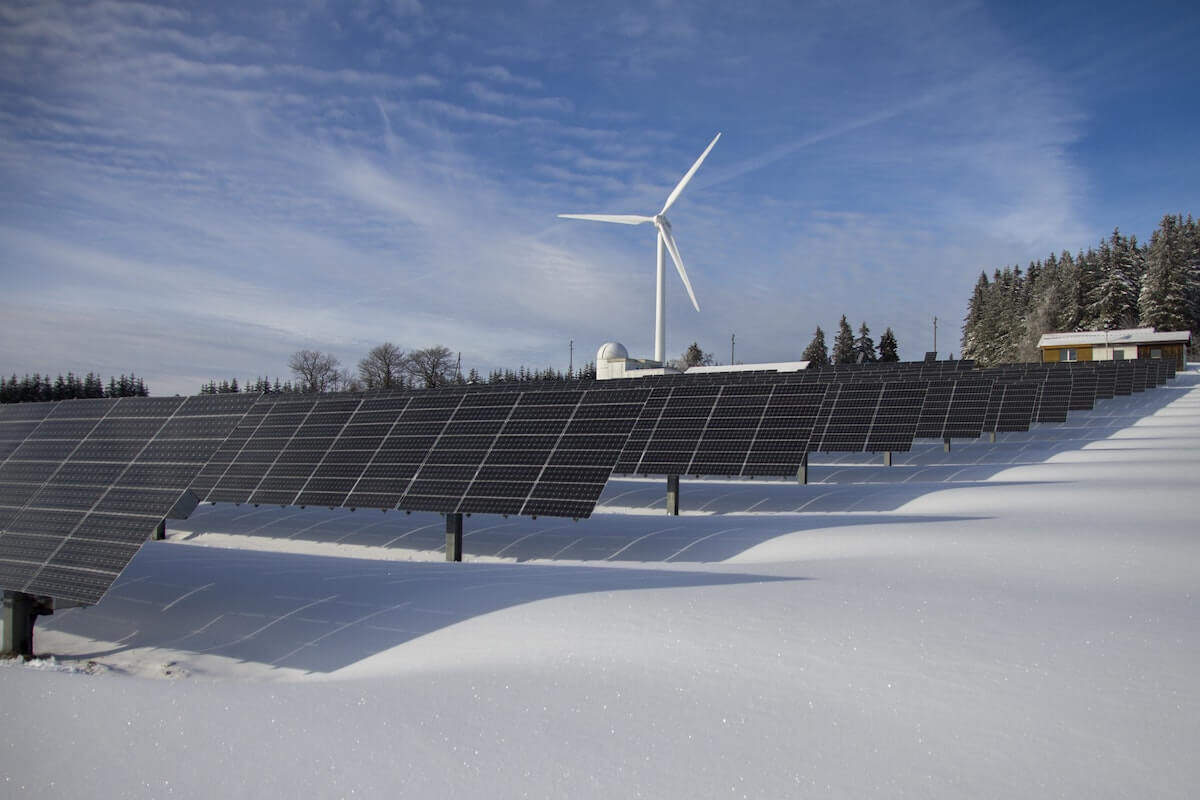 Rows of solar panels and a single wind turbine in a snowy landscape under a blue sky with scattered clouds.