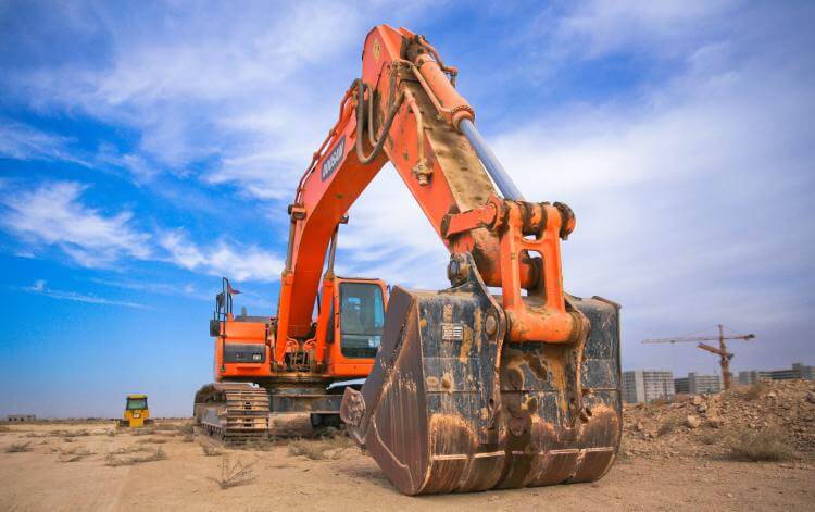 A large orange excavator in a dusty construction site under a clear blue sky, with another excavator in the background and unfinished buildings in the distance.