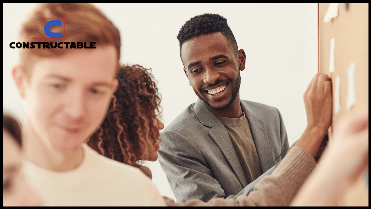A diverse group of professionals in a meeting, with a smiling black man writing the core values on a whiteboard, engaged in an interactive discussion with colleagues. Logo of "constructable" in the corner