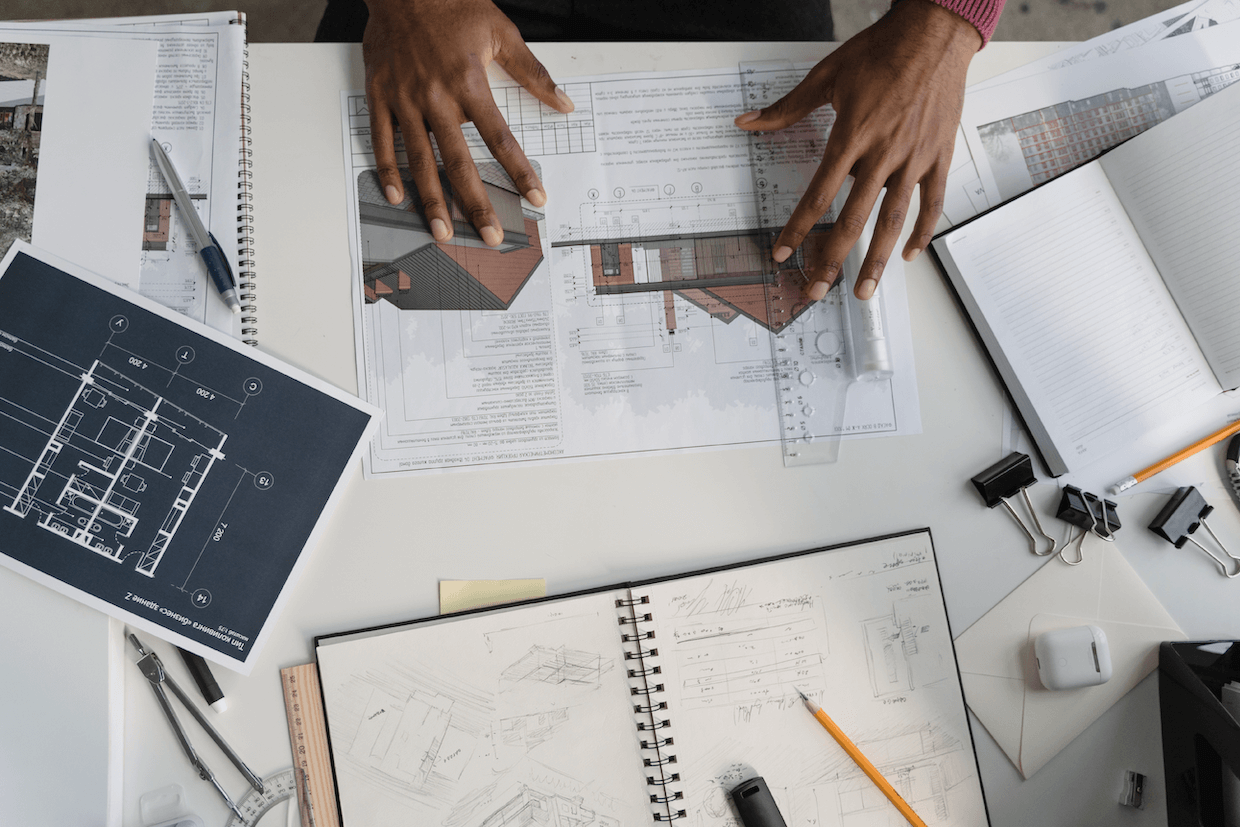 Overhead view of a workspace with architectural drawings and sketches. two hands hold a ruler and a pen over a technical drawing, surrounded by various drafting tools and notebooks.