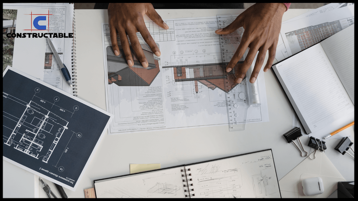 Two people collaborating over architectural designs and drawings on a busy desk, with one using a triangular scale on a blueprint. Various drawing tools and design paperwork are visible.