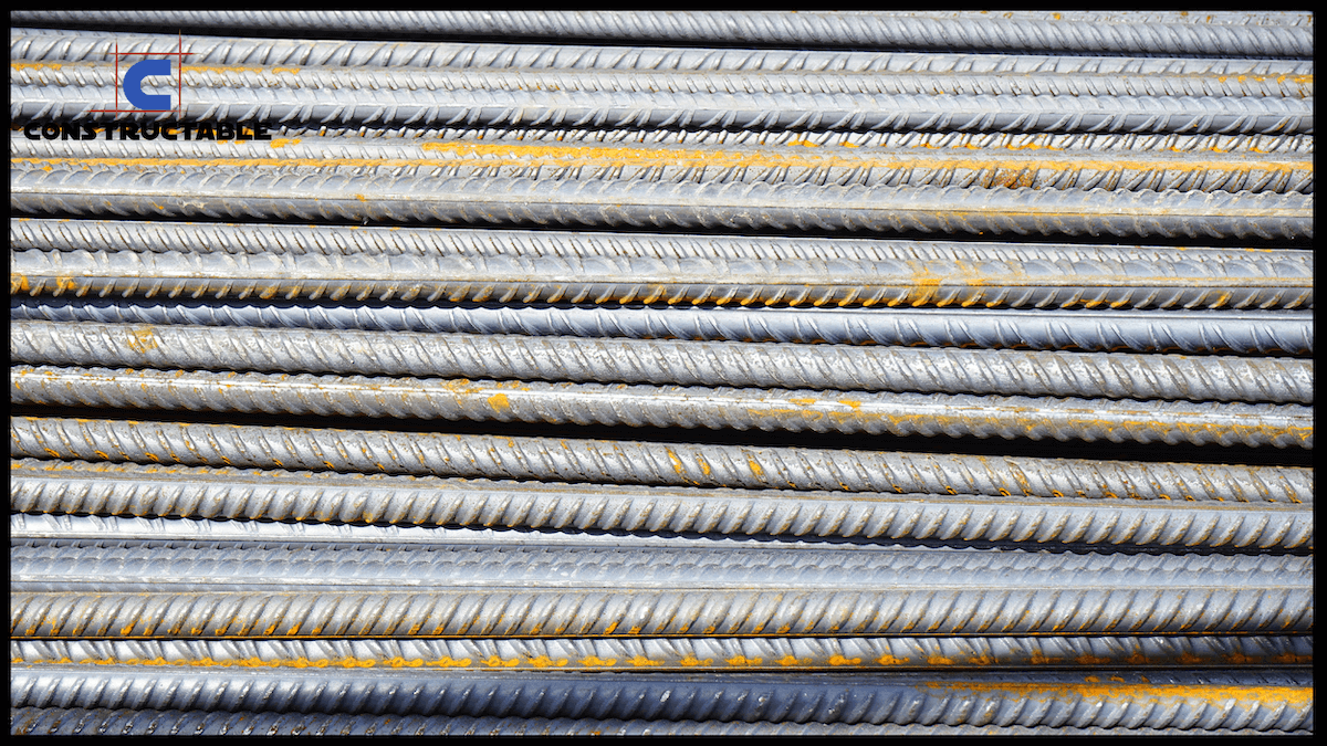 Close-up of stacked steel rebar with visible ridges and slight rust, used in construction reinforcement. The company's design logo is visible in the top left corner.