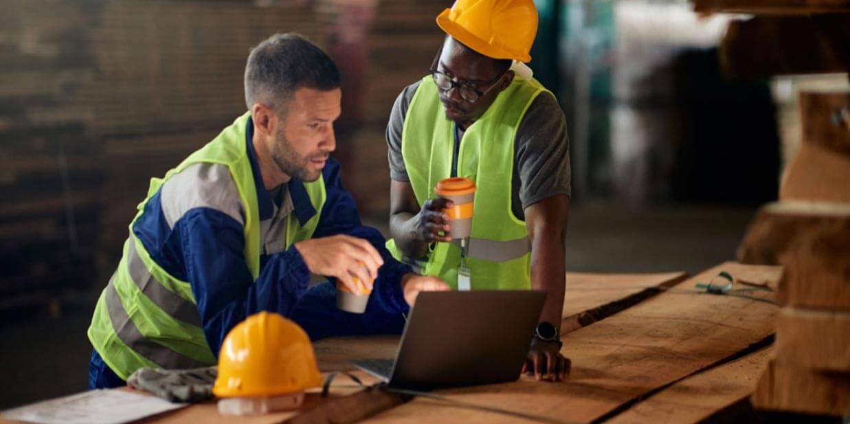 Two construction workers, one holding a coffee cup, looking at a laptop on a wooden table in a warehouse setting, discussing supply chain issues.