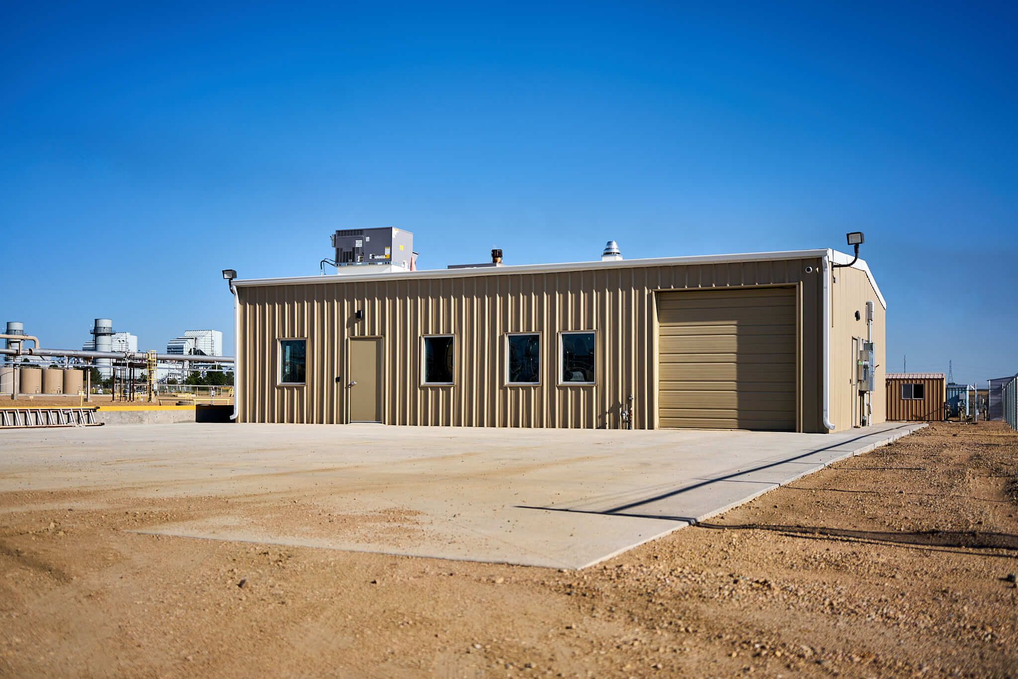 A new tan modular building with several windows, situated on a large concrete pad in an industrial area under a clear blue sky.