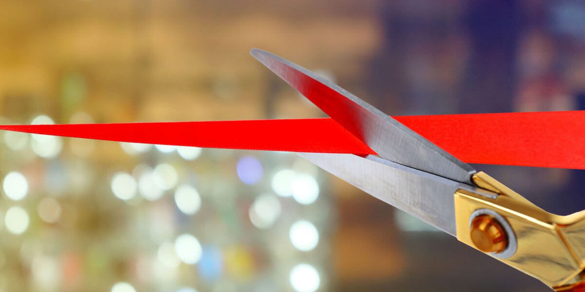 A pair of scissors at a ribbon cutting, slicing through a red ribbon, symbolizing a ceremonial opening, against a blurry background of lights.
