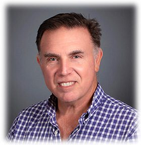 Headshot of a smiling middle-aged man with short dark hair, wearing a blue and white checkered safety shirt against a gray background.