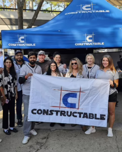 A group of people pose together outside, smiling and holding a banner that reads “CONSTRUCTABLE” in front of a blue Constructable tent with the same logo. Some are wearing matching jerseys, since this was a BBQ competition event.