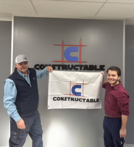Two men stand indoors in front of a wall with a "CONSTRUCTABLE" logo, holding a white company flag featuring the same logo and text. Both are smiling and facing the camera.