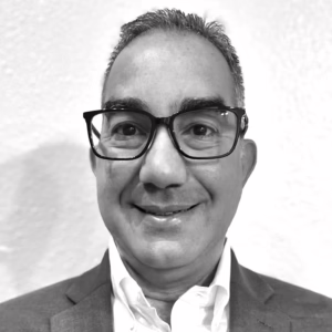 A man with short, dark hair and glasses smiles at the camera. He is wearing a suit jacket and a white collared shirt. The photo is in black and white, with a plain, light-colored background.