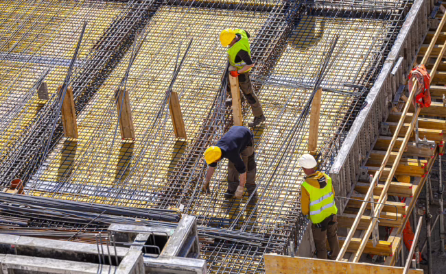 Workers make molds for reinforced concrete from reinforcing bars Three construction workers wearing safety gear and yellow helmets work on a building site, arranging steel reinforcement bars on a large concrete structure under bright daylight.